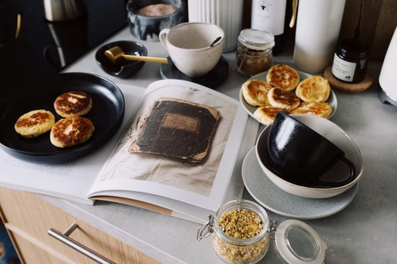 High angle of cookbook and cooked fritters on plates placed on table near utensils and ingredients in cozy kitchen at home